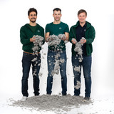 Three men in green jackets and t-shirts stand in front of a white background, dropping grey, fluffy material from their hands onto the ground.